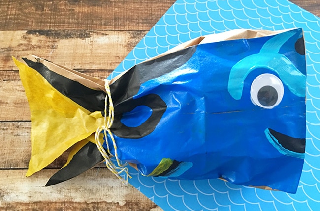 Use your black paint, light blue, and yellow paint to paint Dory's designs on the bag as shown. If you need some guidance, use your sharpie marker to draw on the bag before you start painting. Glue one plastic googly eye onto the top right of the bag. Once your bag is dry, ball up a couple lunch sacks and place them inside your painted bag. Now tie a piece of yellow twine around where the yellow and blue paint meet and you've got a cute little Dory bag.